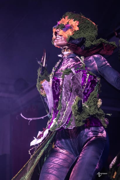 Créature au masque de mousse et de fleurs orange, costume violet à reflets, griffes noires